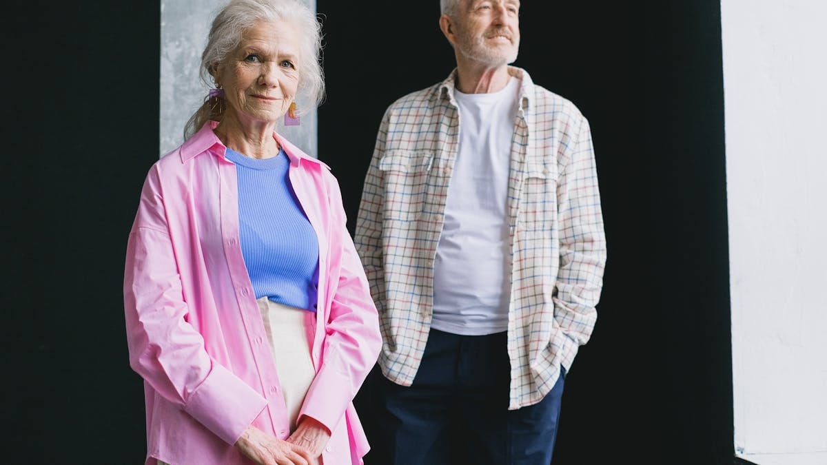 Smiling elderly couple standing indoors wearing colorful casual clothing, exuding warmth and charm.